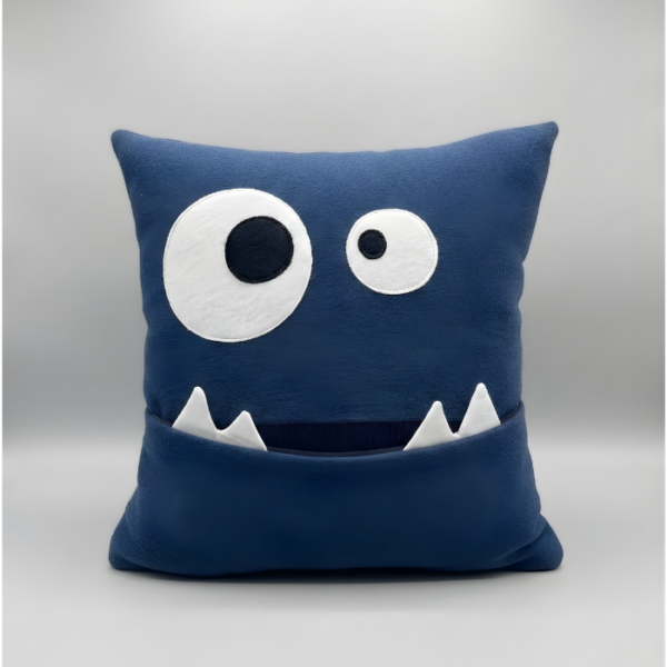 A square, deep blue plush pillow designed as a cute monster face with large, mismatched goofy eyes and a wide mouth that functions as a pocket with two white felt teeth protruding.
