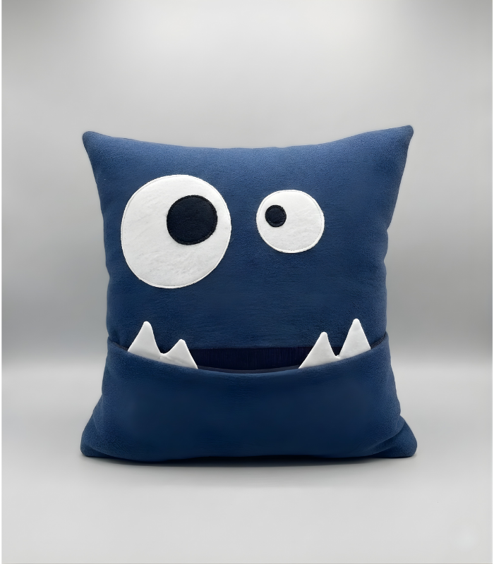 A square, deep blue plush pillow designed as a cute monster face with large, mismatched goofy eyes and a wide mouth that functions as a pocket with two white felt teeth protruding.