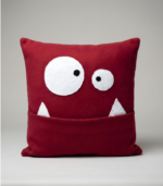 A deep red, square plush pillow designed as a cute monster face. It features two mismatched, surprised white eyes and two white felt teeth. The wide mouth functions as a deep pocket for storing items.