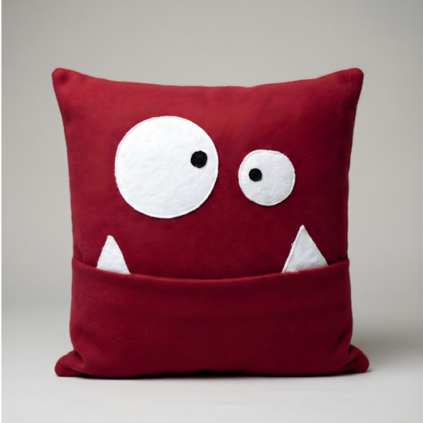 A deep red, square plush pillow designed as a cute monster face. It features two mismatched, surprised white eyes and two white felt teeth. The wide mouth functions as a deep pocket for storing items.